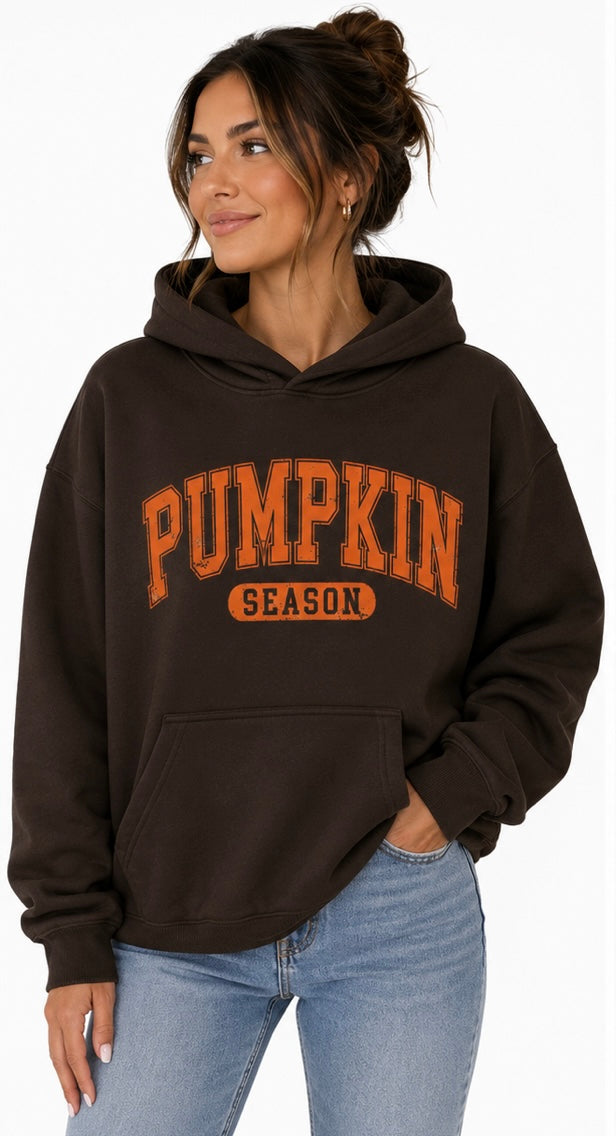 A woman wearing a dark brown pullover hoodie with 'PUMPKIN SEASON' printed in orange on the front, paired with light blue jeans, against a plain white background.