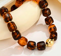 A beaded stretch bracelet made of glossy, rounded brown tiger's eye beads with small gold spacer rings and a polished gold ball accent, displayed on a cream-colored curved stand against a white background.