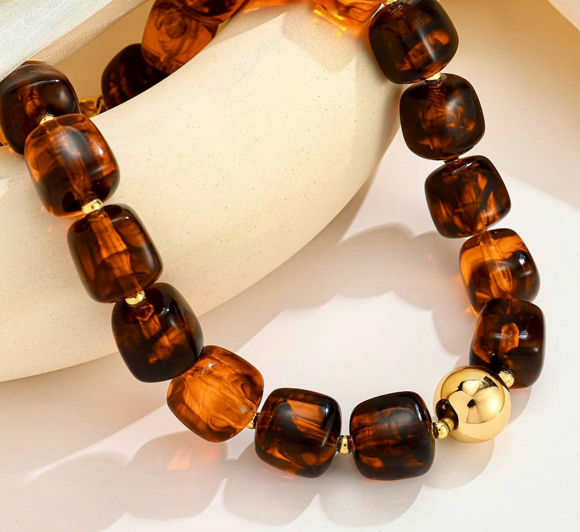 A beaded stretch bracelet made of glossy, rounded brown tiger's eye beads with small gold spacer rings and a polished gold ball accent, displayed on a cream-colored curved stand against a white background.