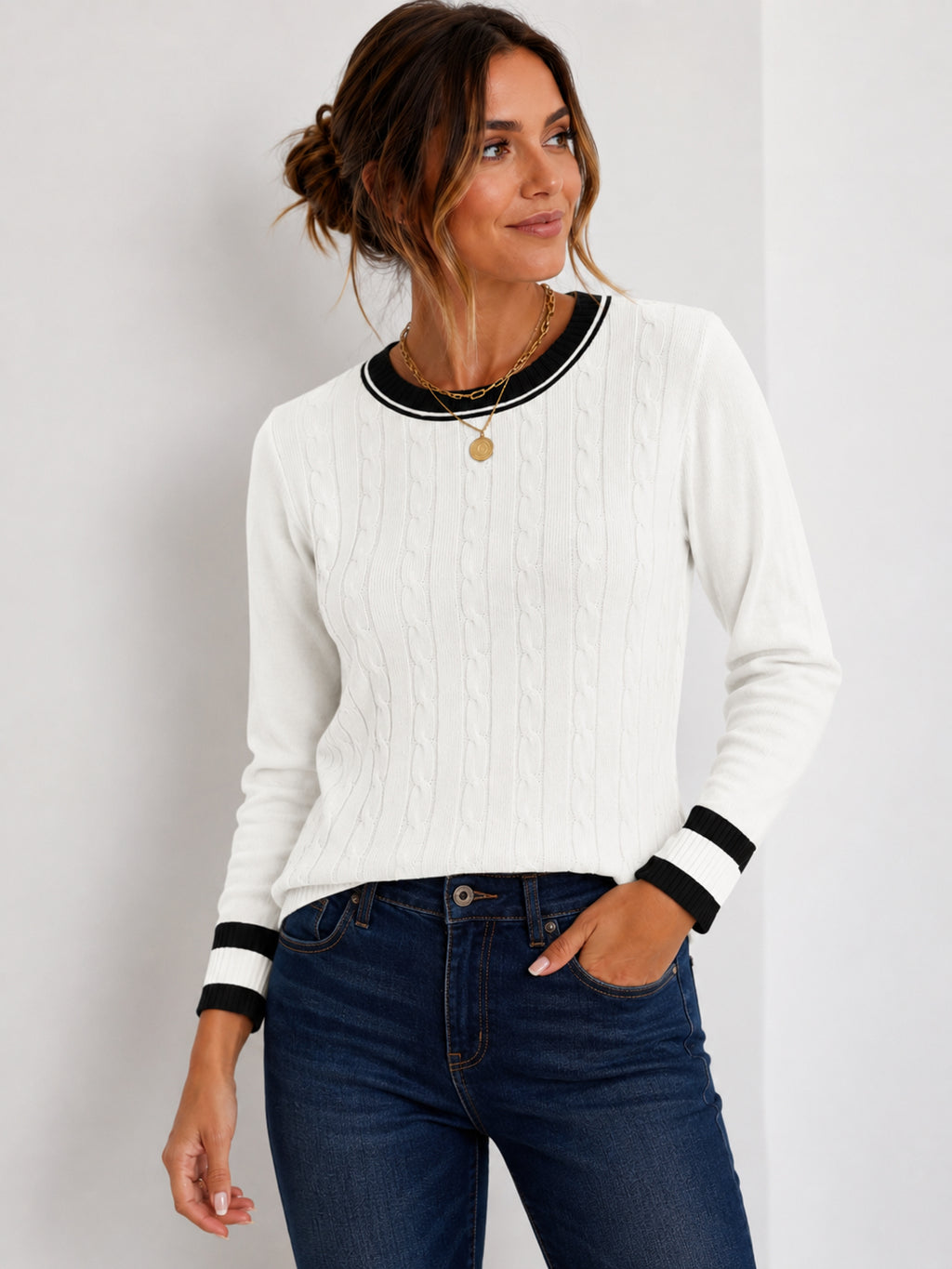 A woman wearing a white cable-knit long-sleeve sweater with black trim at the crew neck and cuffs, paired with dark blue jeans, against a light gray studio background.