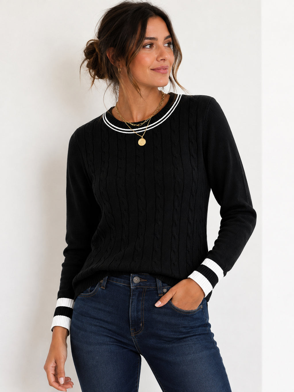A woman wearing a black cable-knit sweater with white striped trim at the crew neck and cuffs, paired with dark blue jeans, against a plain white background.