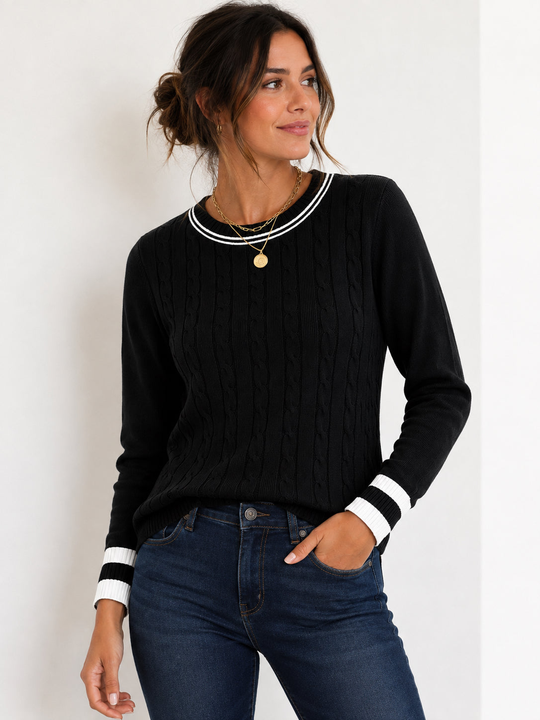 A woman wearing a black cable-knit sweater with white striped trim at the crew neck and cuffs, paired with dark blue jeans, against a plain white background.