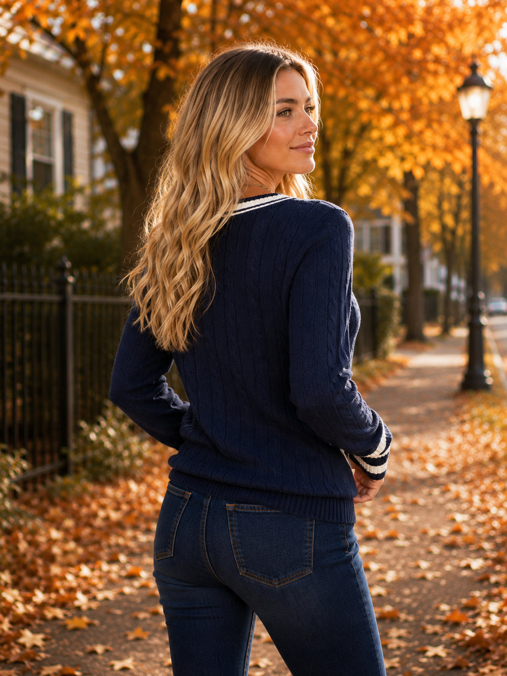 A woman standing on an autumn tree-lined path, wearing a navy blue cable-knit sweater with white striped trim and dark blue jeans.