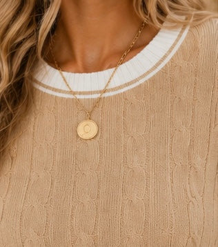 A close-up of a woman wearing a gold chain necklace with a round initial pendant, styled with a beige cable-knit sweater and a white ribbed crew neck.