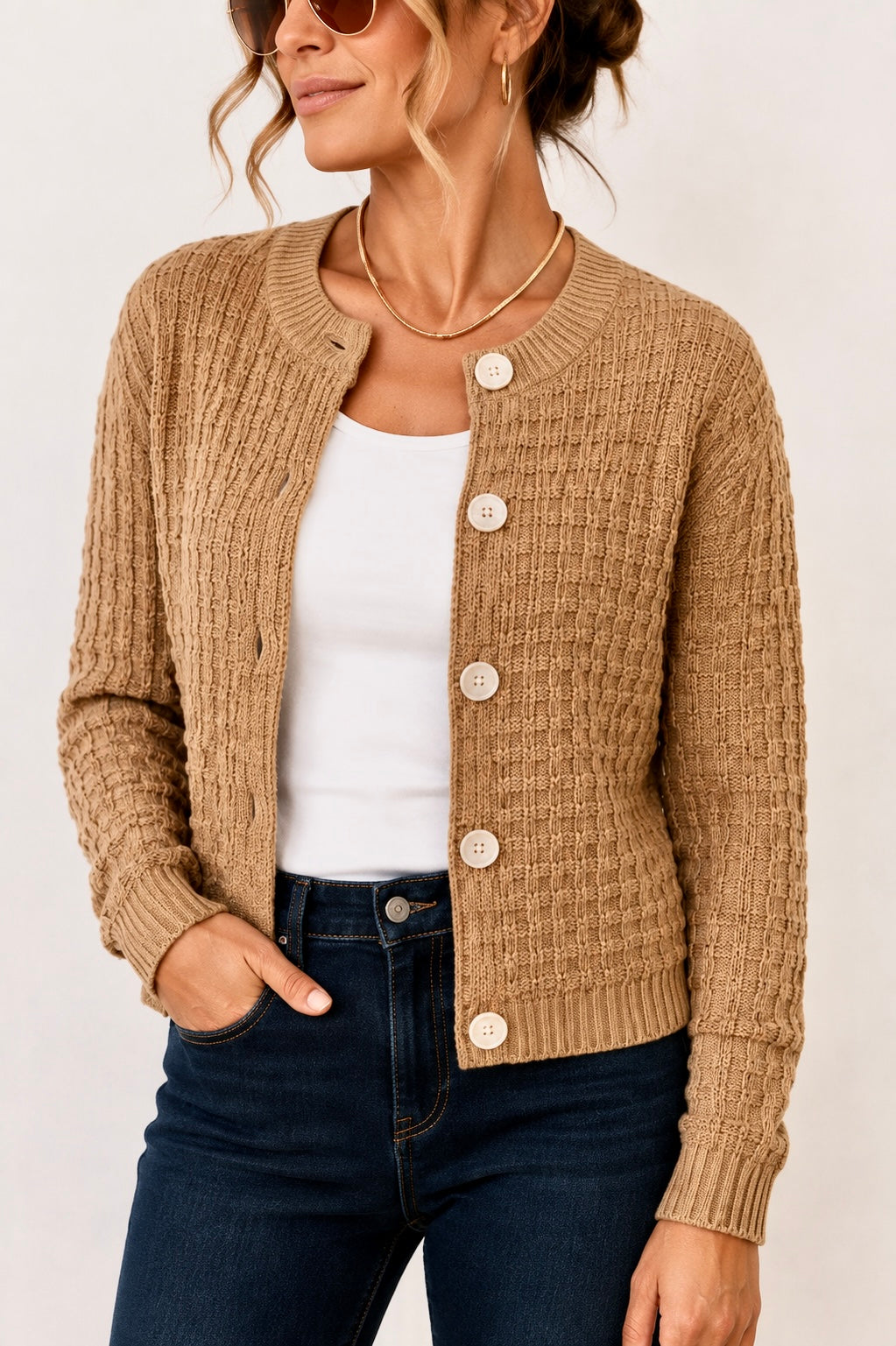 A woman wearing a tan cable-knit button-front cardigan over a white tank top and dark blue jeans, photographed against a plain light background.