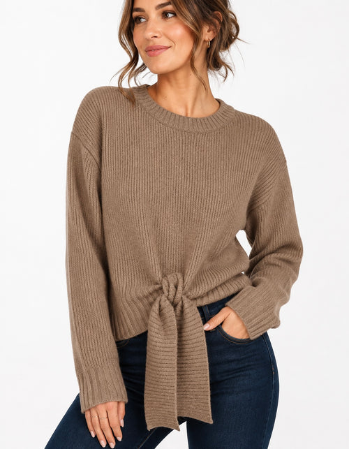 A woman wearing a taupe brown ribbed knit sweater with a front knot detail and dark blue jeans, photographed against a clean white background.
