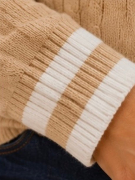 Close-up of a beige cable-knit sweater sleeve with two thick white horizontal stripes at the cuff, worn over dark blue jeans.