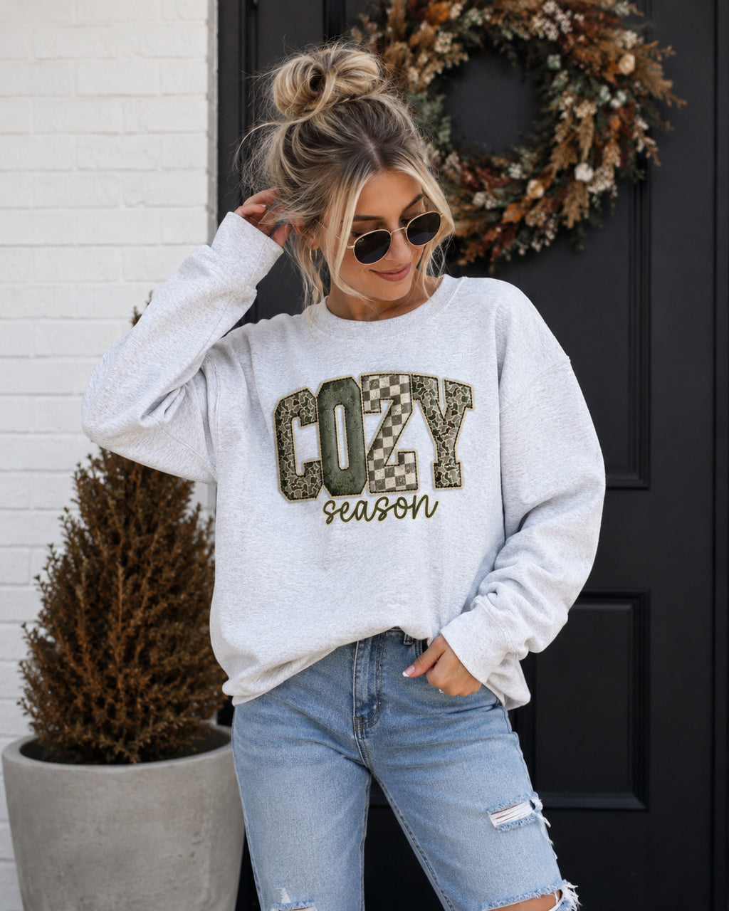 A woman wearing a light gray crewneck sweatshirt with 'COZY season' printed on the front, paired with light blue jeans, standing in front of a black door with a wreath.