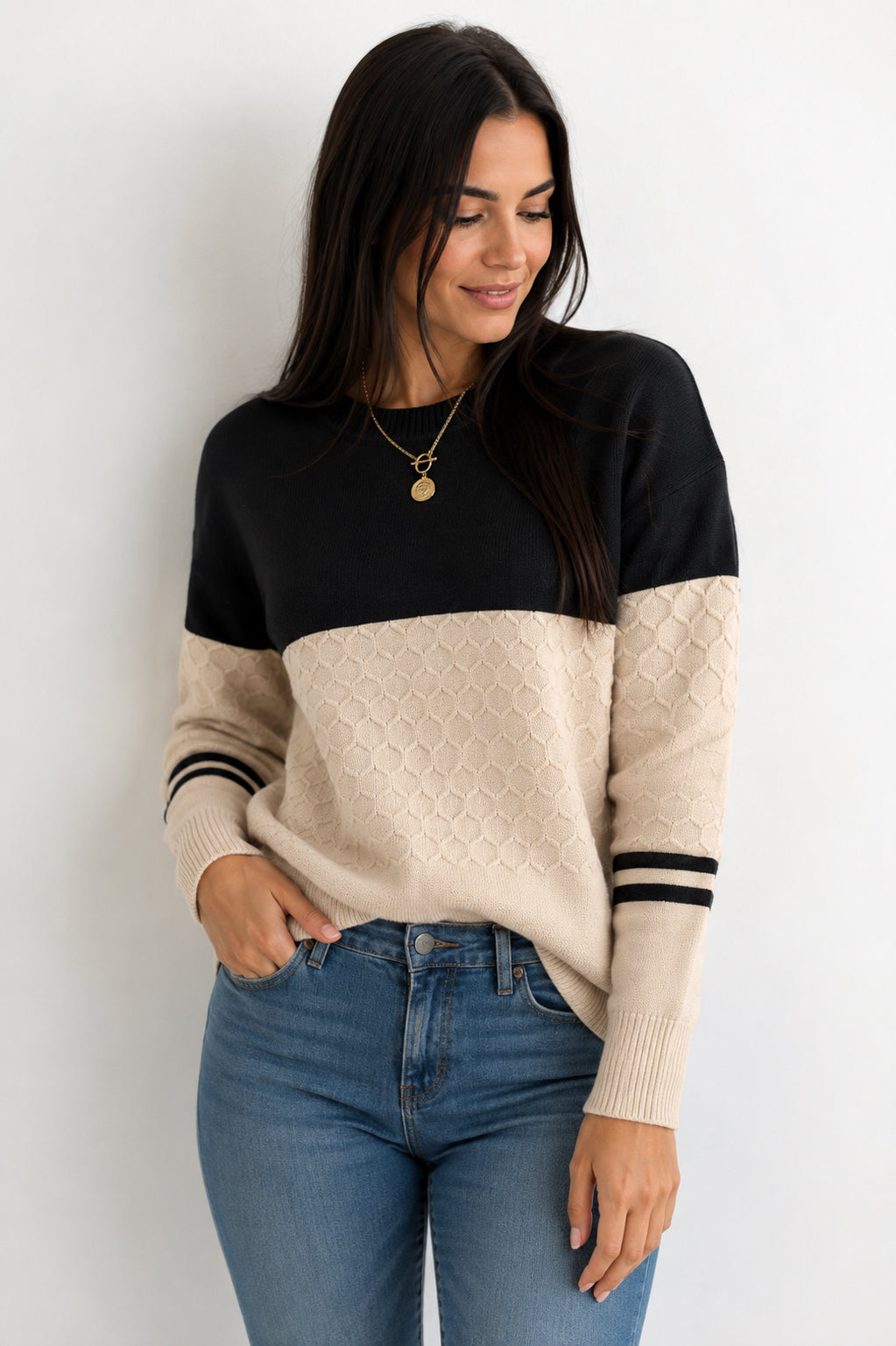 A woman wearing a color-block knit sweater with a black upper section and a textured beige lower section and sleeves, paired with blue jeans against a plain white background.