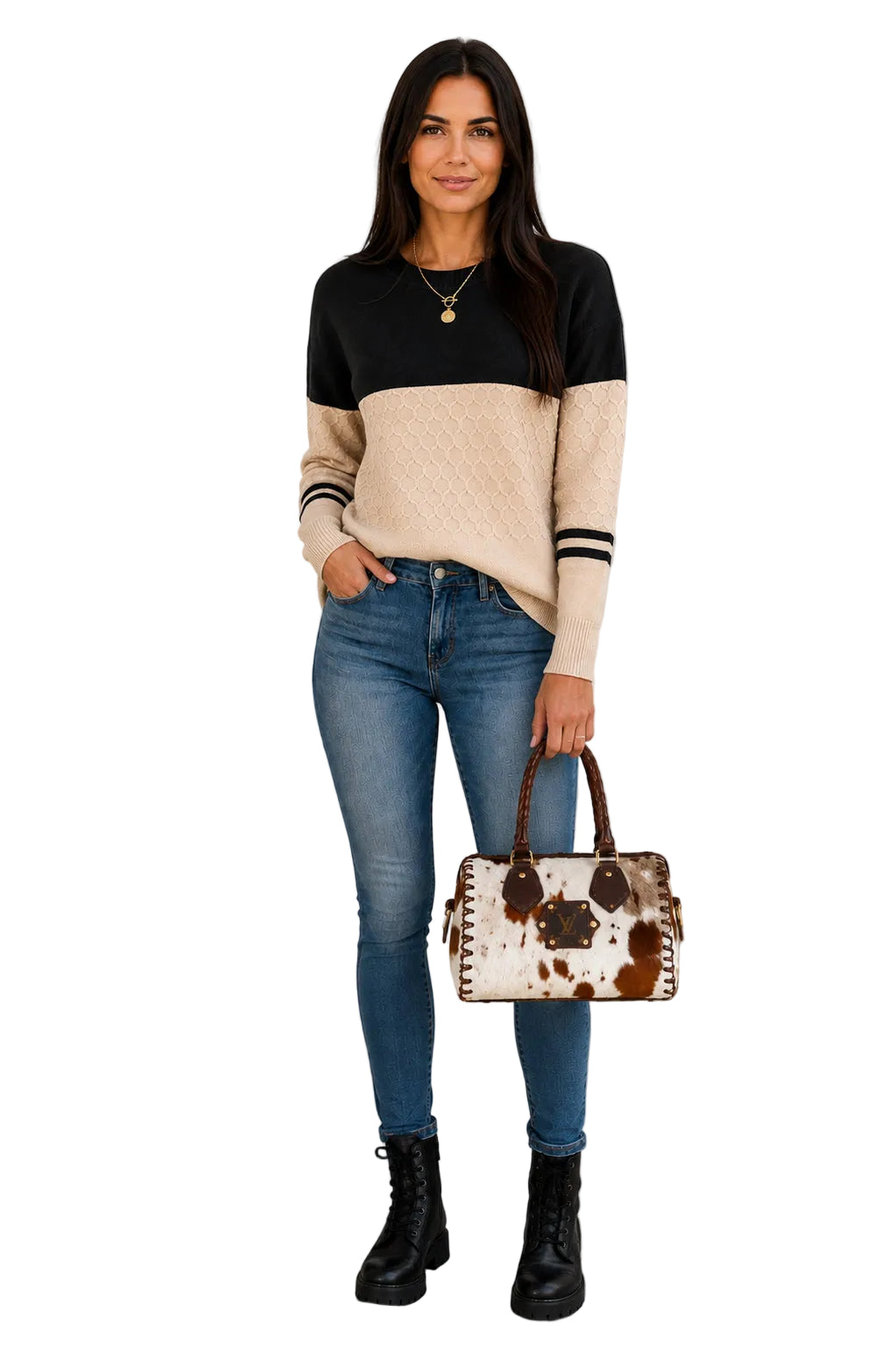 A woman stands outdoors wearing a color-block knit sweater with black and beige sections, blue skinny jeans, black lace-up boots, and holding a cow-print handbag.