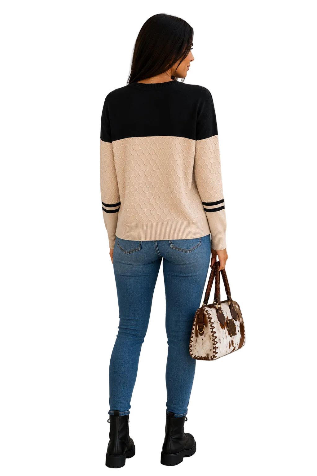 A woman viewed from behind wearing a color-block knit sweater with a black top section, beige middle band, and beige sleeves with black stripes. She is wearing blue skinny jeans and black boots, holding a cow-print handbag with a white background.