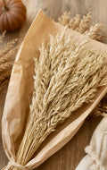 A bundle of dried wheat stalks wrapped in brown kraft paper, arranged on a rustic wooden surface with a small pumpkin and neutral fabric in the background.