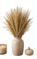 A tall textured cream ceramic vase filled with dried wheat or ornamental grass stems, displayed on a wooden surface with a lit candle, a small white pumpkin, and an orange pumpkin with white background.