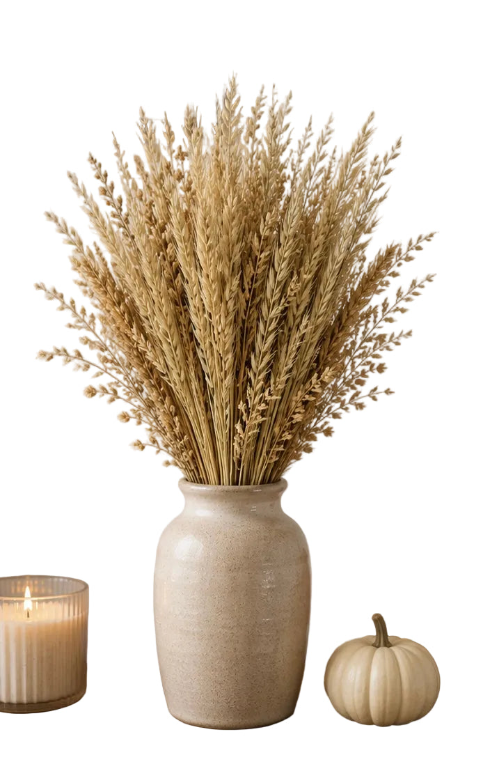 A tall textured cream ceramic vase filled with dried wheat or ornamental grass stems, displayed on a wooden surface with a lit candle, a small white pumpkin, and an orange pumpkin with white background.