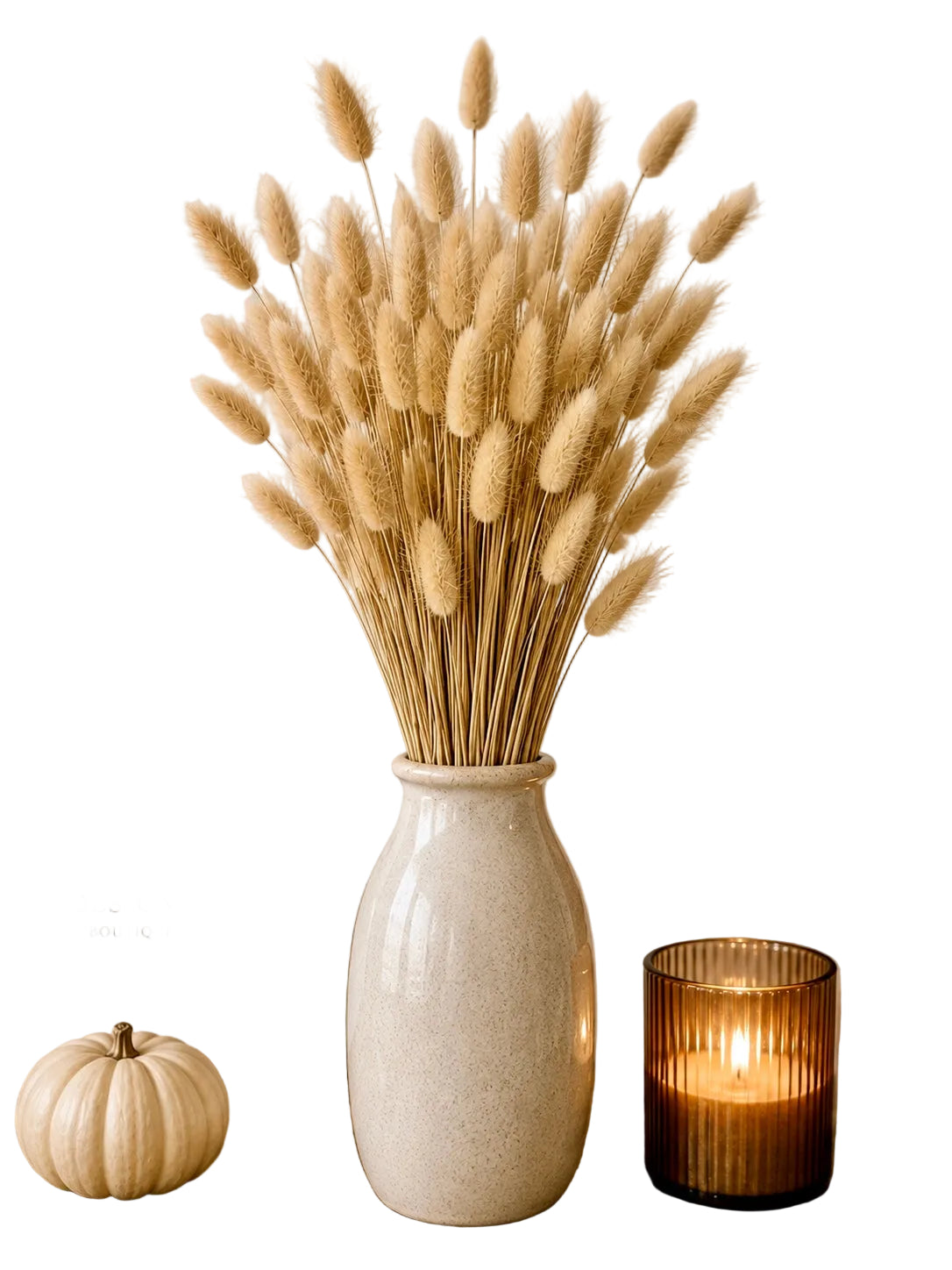 A tall cream ceramic vase filled with dried bunny tail grass, styled on a wooden tabletop with a small cream pumpkin, a framed print reading 'heartfelt DESIGNS BOUTIQUE', a lit amber glass candle holder, and a potted autumn leaf arrangement against a white background.