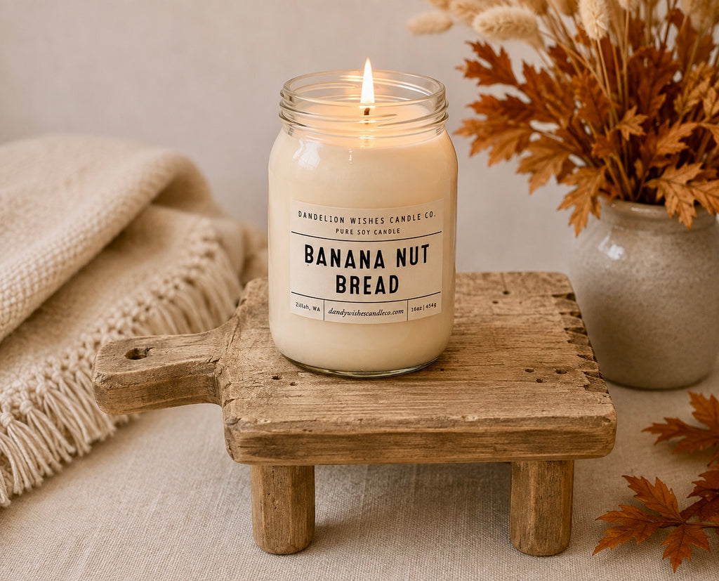 A lit candle in a clear glass jar sits on a rustic wooden stand. The label reads 'Banana Nut Bread' and shows the brand 'Bansellon Wishes Candle Co.