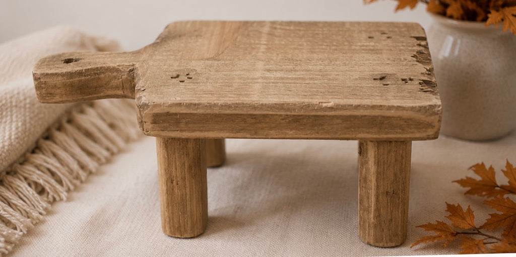 A rustic wooden tabletop stool with a thick rectangular top, four short legs, and a handle on the left side, styled on a cream fabric with a throw blanket and dried leaves.