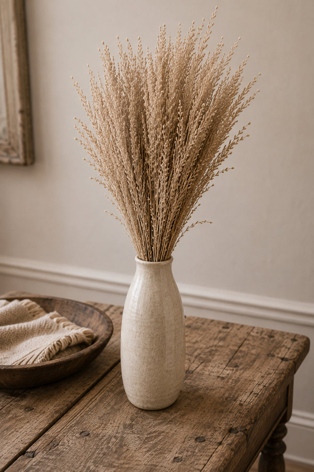 A tall textured cream ceramic vase filled with dried beige grasses, displayed on a rustic wooden table next to a woven bowl with a neutral throw blanket in a softly lit room.