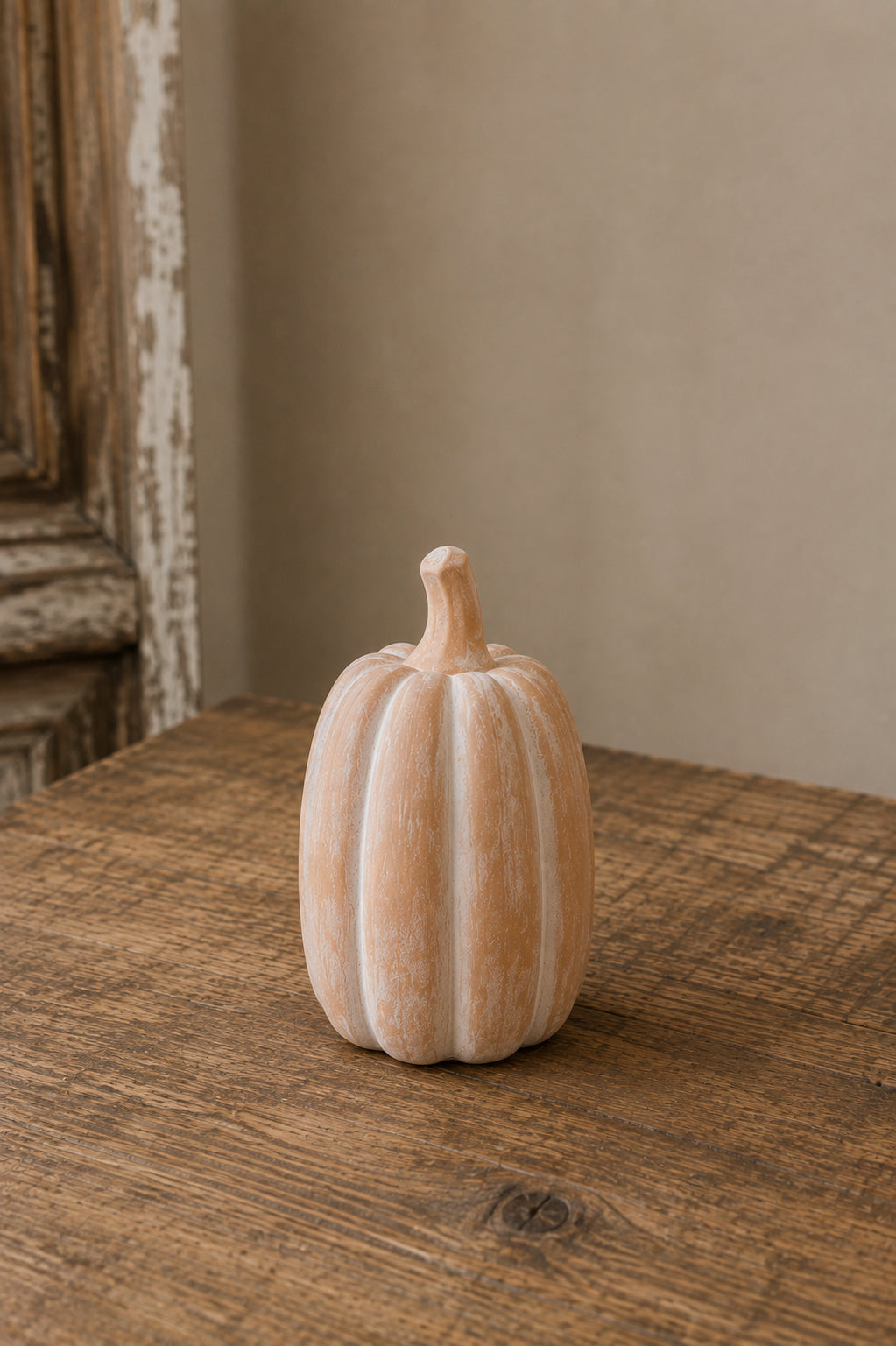 A small ribbed decorative pumpkin in a warm off-white or cream tone, placed on a rustic wooden tabletop against a soft beige wall.