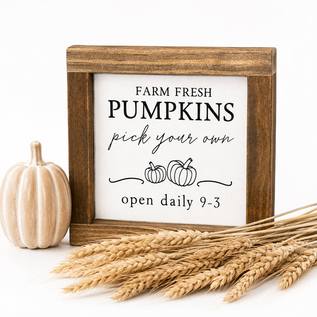 A square wooden sign with a rustic brown frame and a white face reading 'FARM FRESH PUMPKINS pick your own open daily 9-3' with small pumpkin illustrations. The sign is styled on a white background with a small decorative pumpkin and wheat stalks.