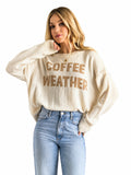 A woman wearing a cream-colored knit sweater with 'COFFEE WEATHER' printed in brown across the chest, paired with light blue jeans, against a plain white background.