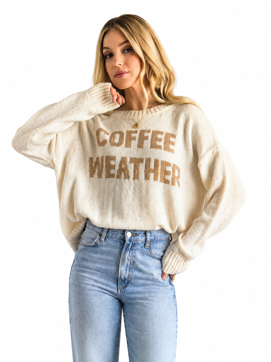 A woman wearing a cream-colored knit sweater with 'COFFEE WEATHER' printed in brown across the chest, paired with light blue jeans, against a plain white background.