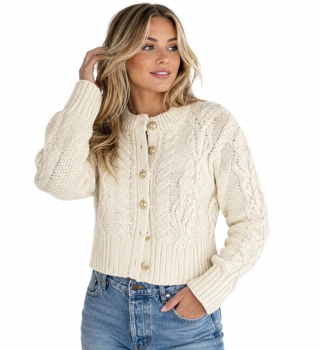 A woman wearing a cream cable-knit cardigan sweater with gold buttons, paired with blue jeans, against a solid white background.
