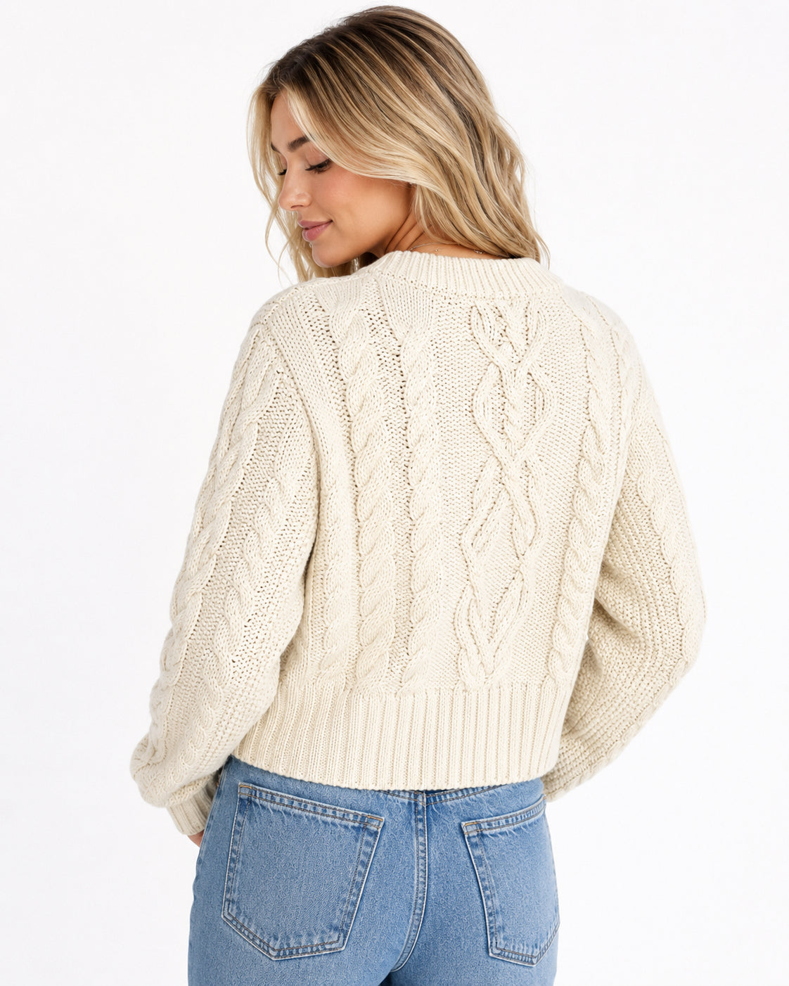 A woman shown from the back wearing a cream cable-knit cropped sweater and light blue jeans against a plain white studio background.