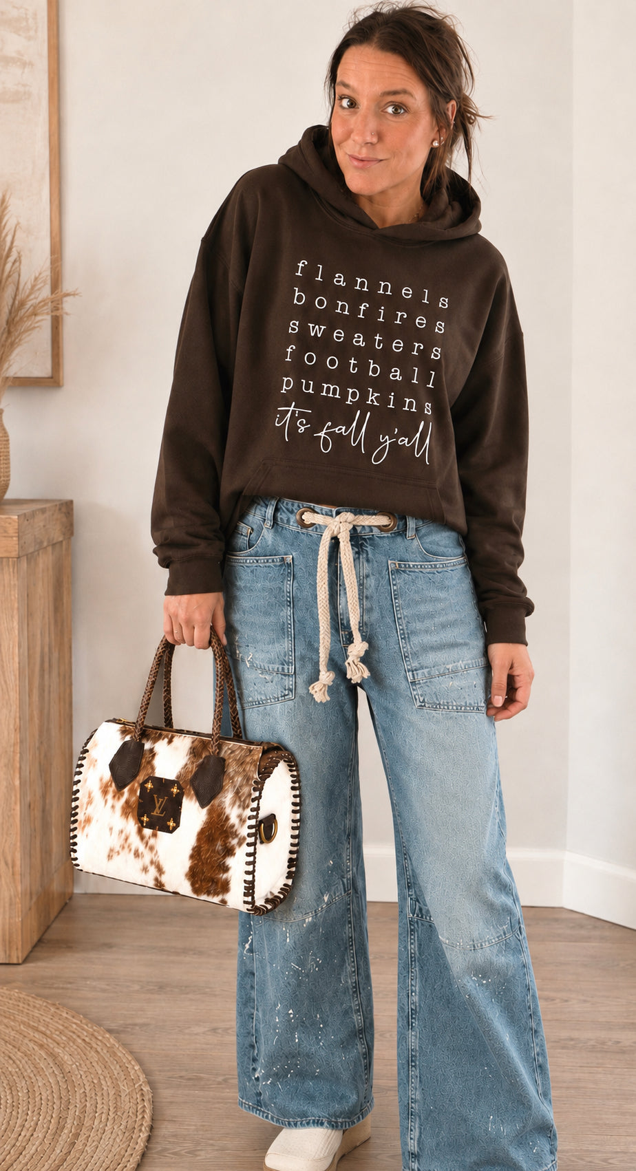 A woman stands indoors wearing a dark brown hoodie with white text listing fall activities, paired with wide-leg light blue jeans and holding a cow-print handbag.