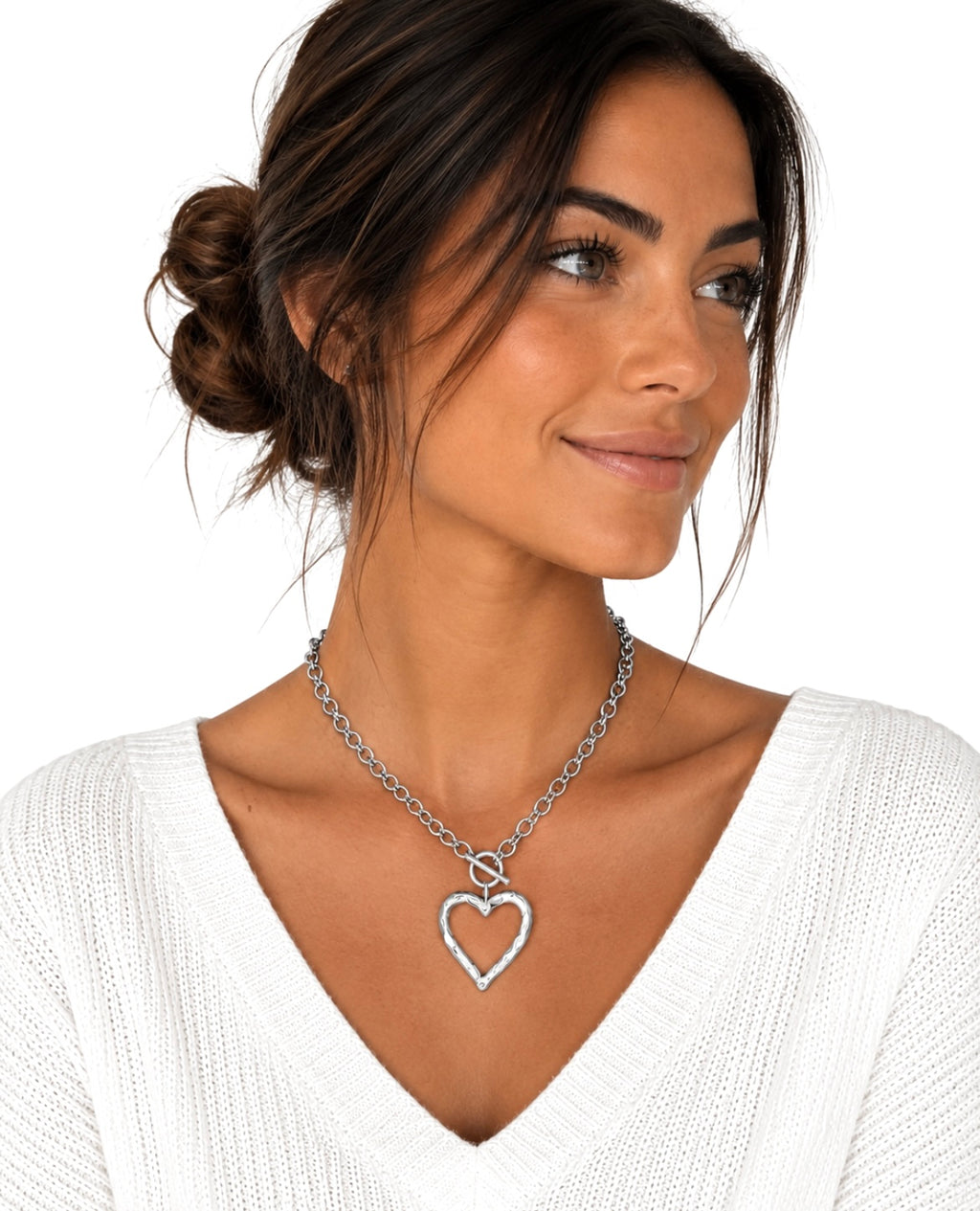 A woman with dark hair in a bun wearing a silver heart-shaped pendant necklace over a white knit top, photographed against a plain white background.