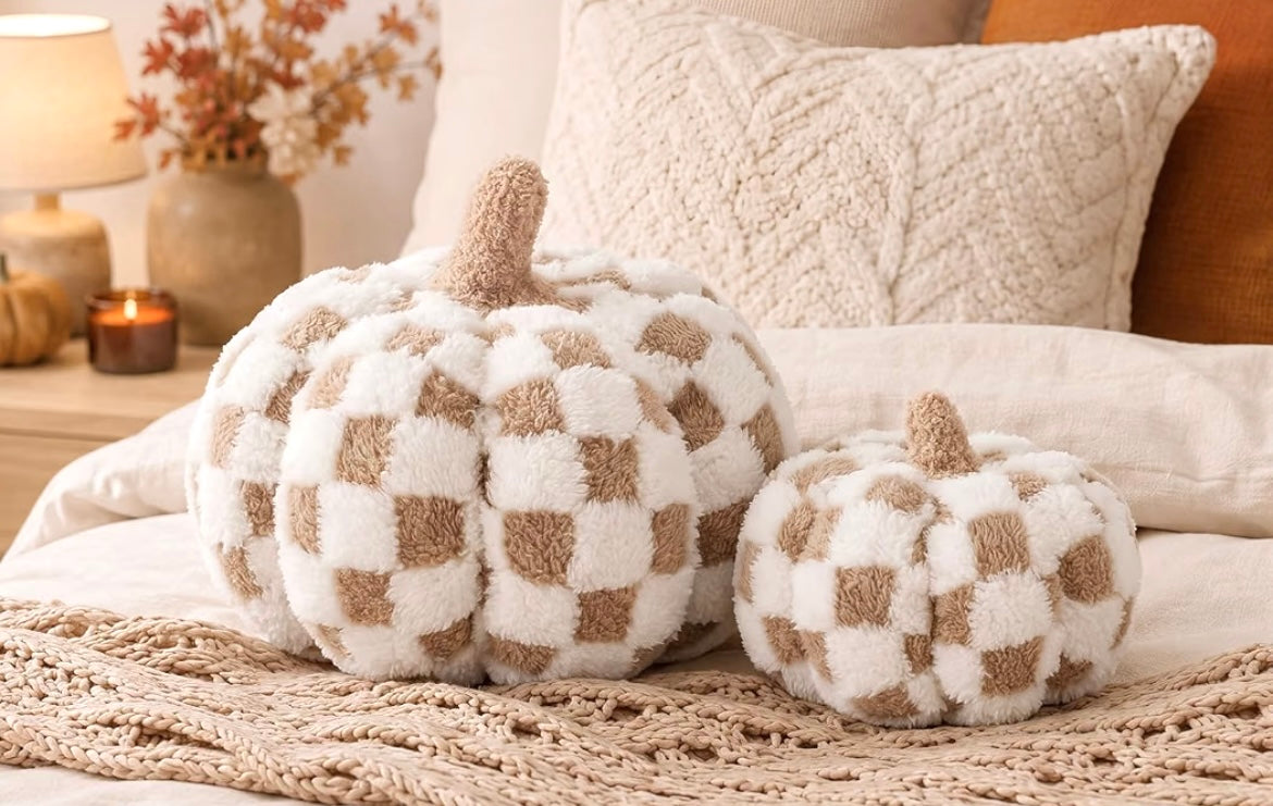 Two plush decorative pumpkins in a cream and tan checkerboard pattern, styled on a bed with cozy pillows and a knit blanket in a warm autumn bedroom setting.