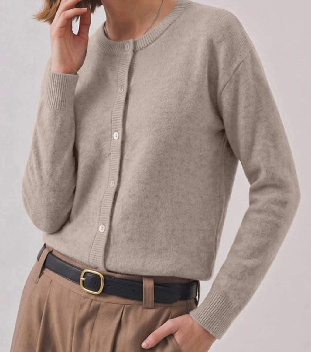 A person wearing a light taupe knit cardigan sweater with white buttons, paired with brown trousers and a black belt with a gold buckle, against a plain light gray background.
