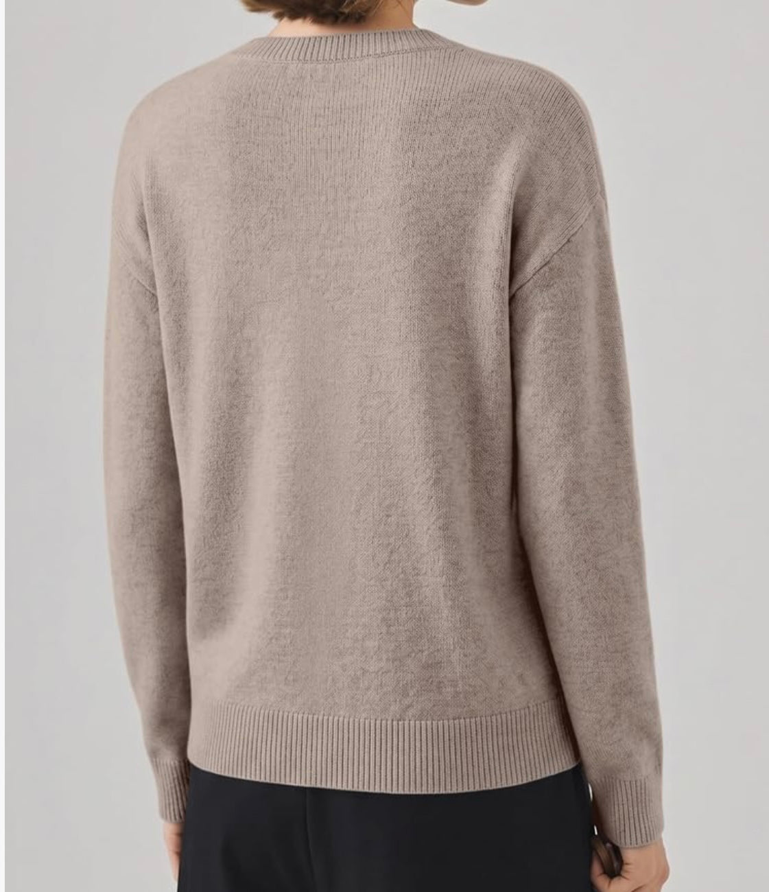 A person shown from the back wearing a light taupe knit sweater with ribbed collar, cuffs, and hem, paired with dark pants against a plain light gray studio background.