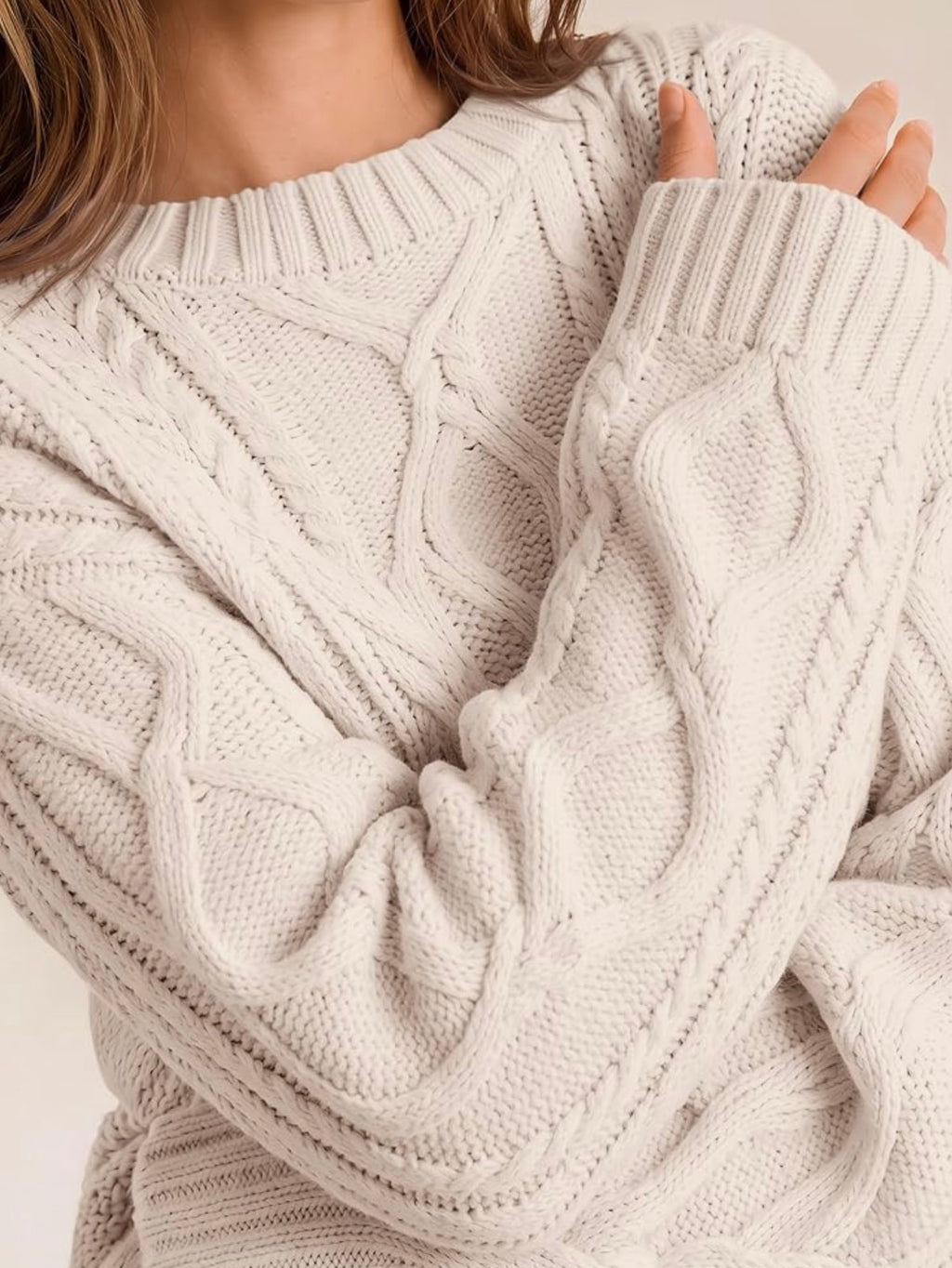 Close-up of a person wearing a chunky cable-knit sweater in a warm off-white or cream tone, showing the detailed knit texture and ribbed collar and cuffs.