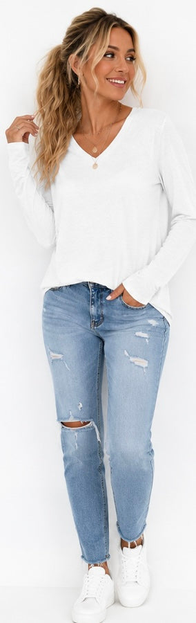 A woman modeling a white long-sleeve V-neck top and light-wash distressed skinny jeans with ripped knees, paired with white sneakers, against a plain white background.
