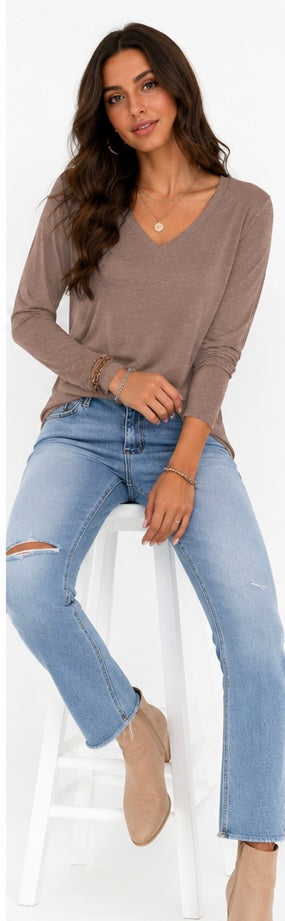 A woman wearing a taupe brown long-sleeve V-neck top, light blue distressed jeans, and beige ankle boots, sitting on a white stool against a white background.