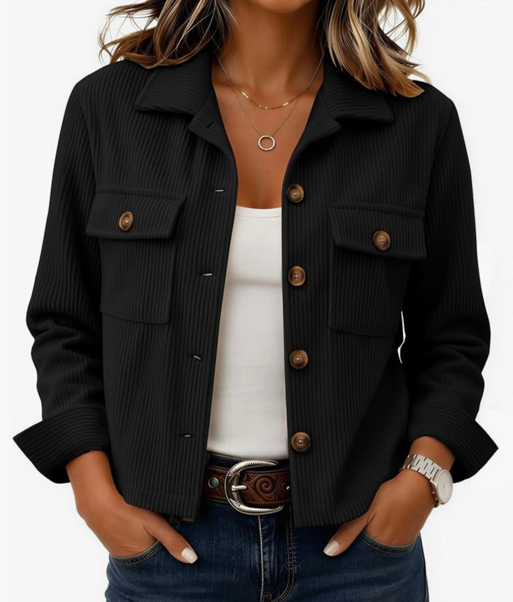 A woman wearing a black ribbed button-front jacket with flap chest pockets and brown buttons, layered over a white tank top and blue jeans.
