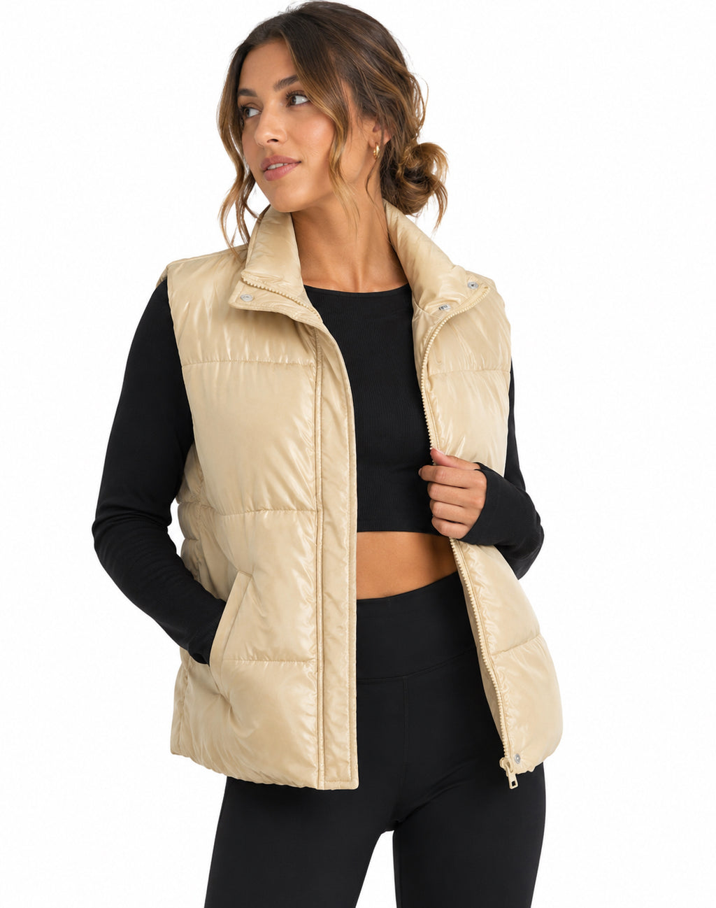 A woman wearing a light beige puffer vest over a black long-sleeve crop top and black leggings, photographed against a plain white studio background.