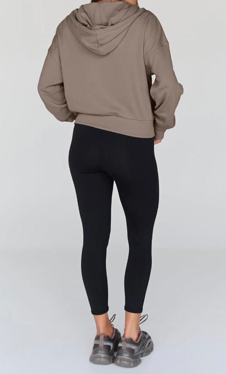 A person shown from the back wearing a cropped taupe-brown hooded sweatshirt, black leggings, and gray sneakers against a light gray studio background.