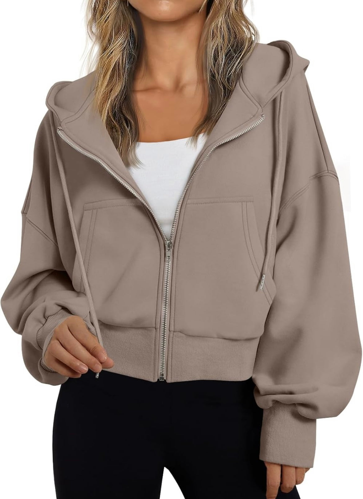 A woman wearing a taupe-brown cropped zip-up hoodie with a drawstring hood and front pockets, layered over a white top and black pants, against a plain white background.