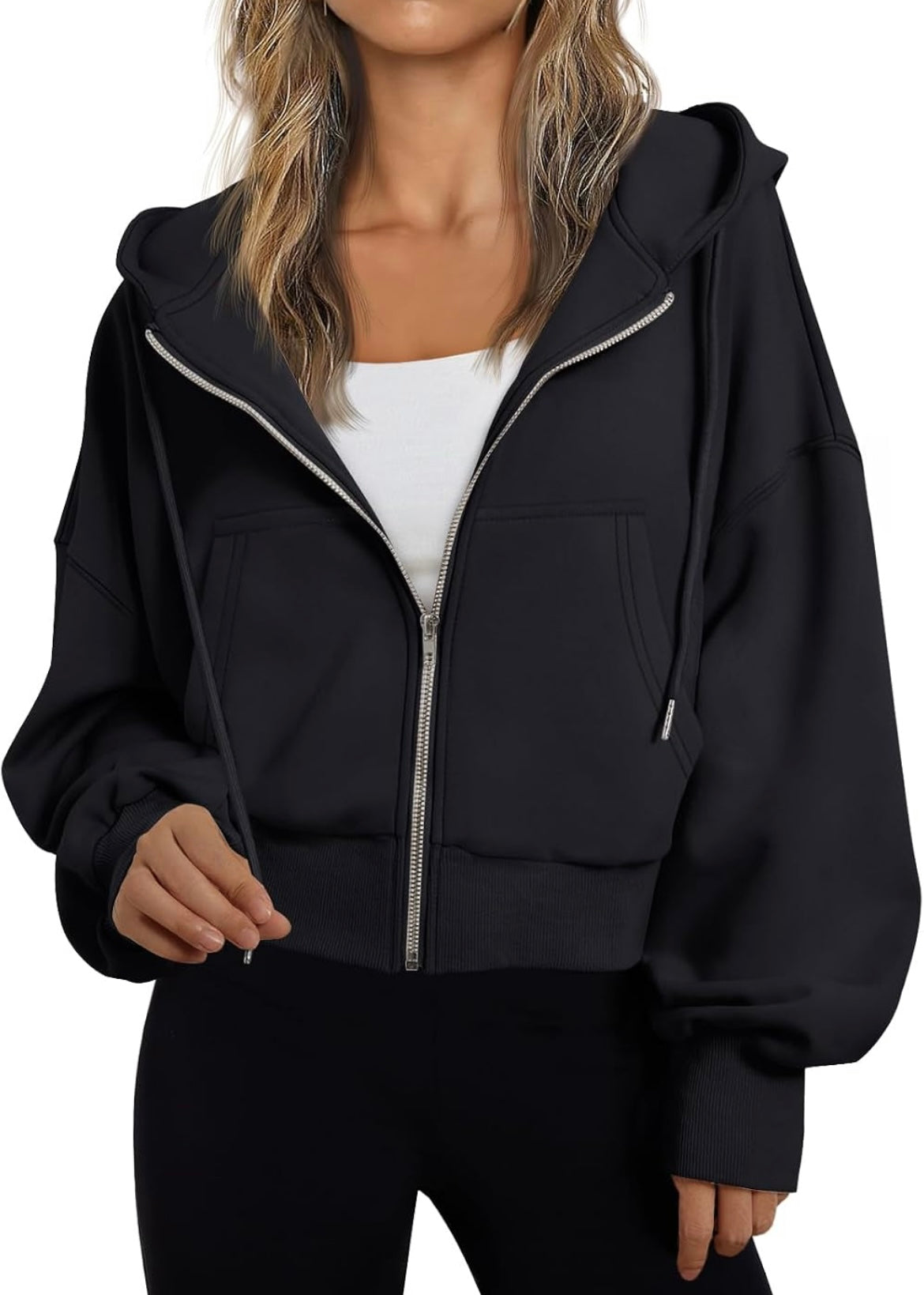 A woman wearing a black cropped zip-up hoodie with silver zipper accents over a white top and black pants, shown against a plain white background.