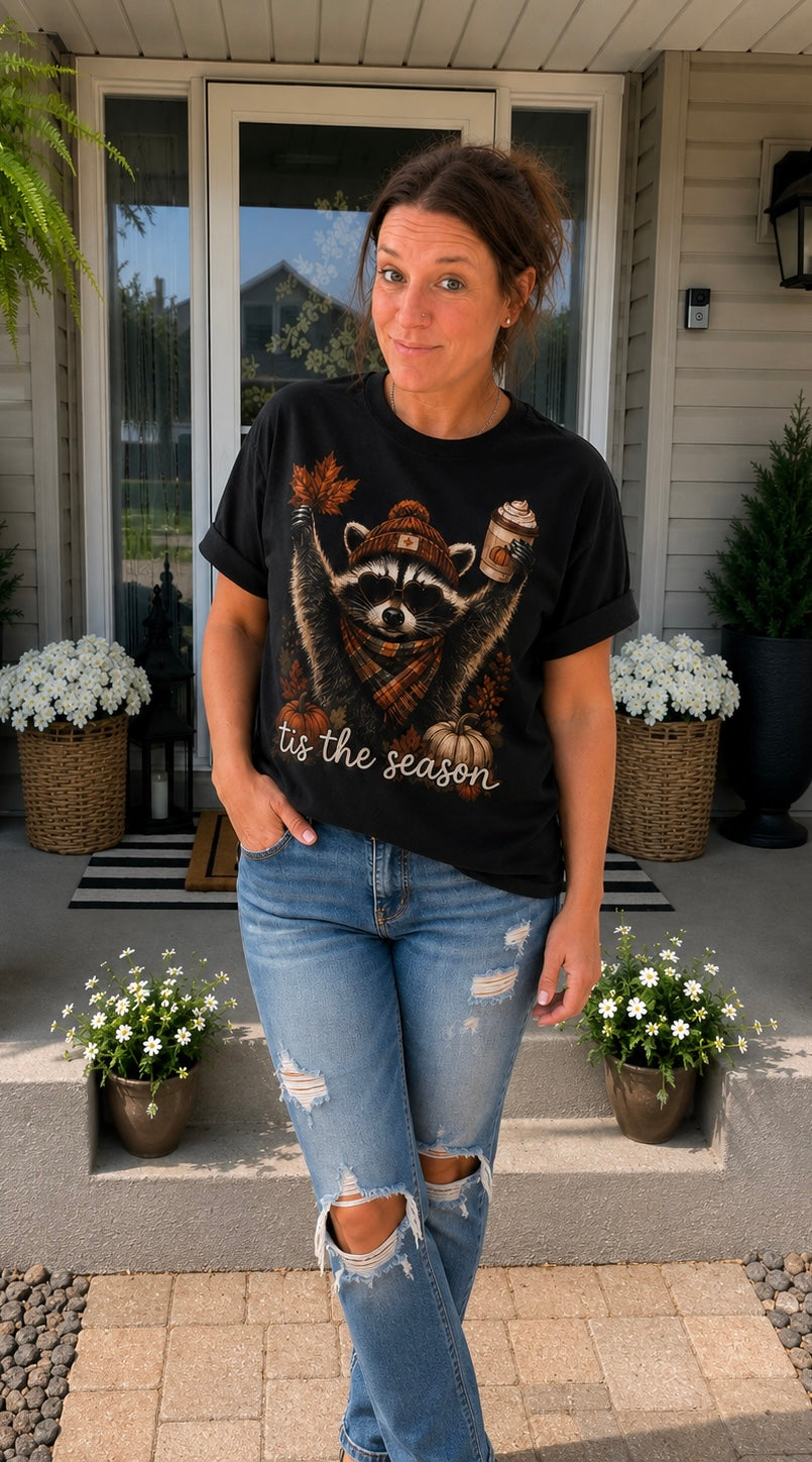 A woman stands on a front porch wearing a black graphic t-shirt with a raccoon wearing a Santa hat and the text 'its the season', paired with distressed blue jeans.