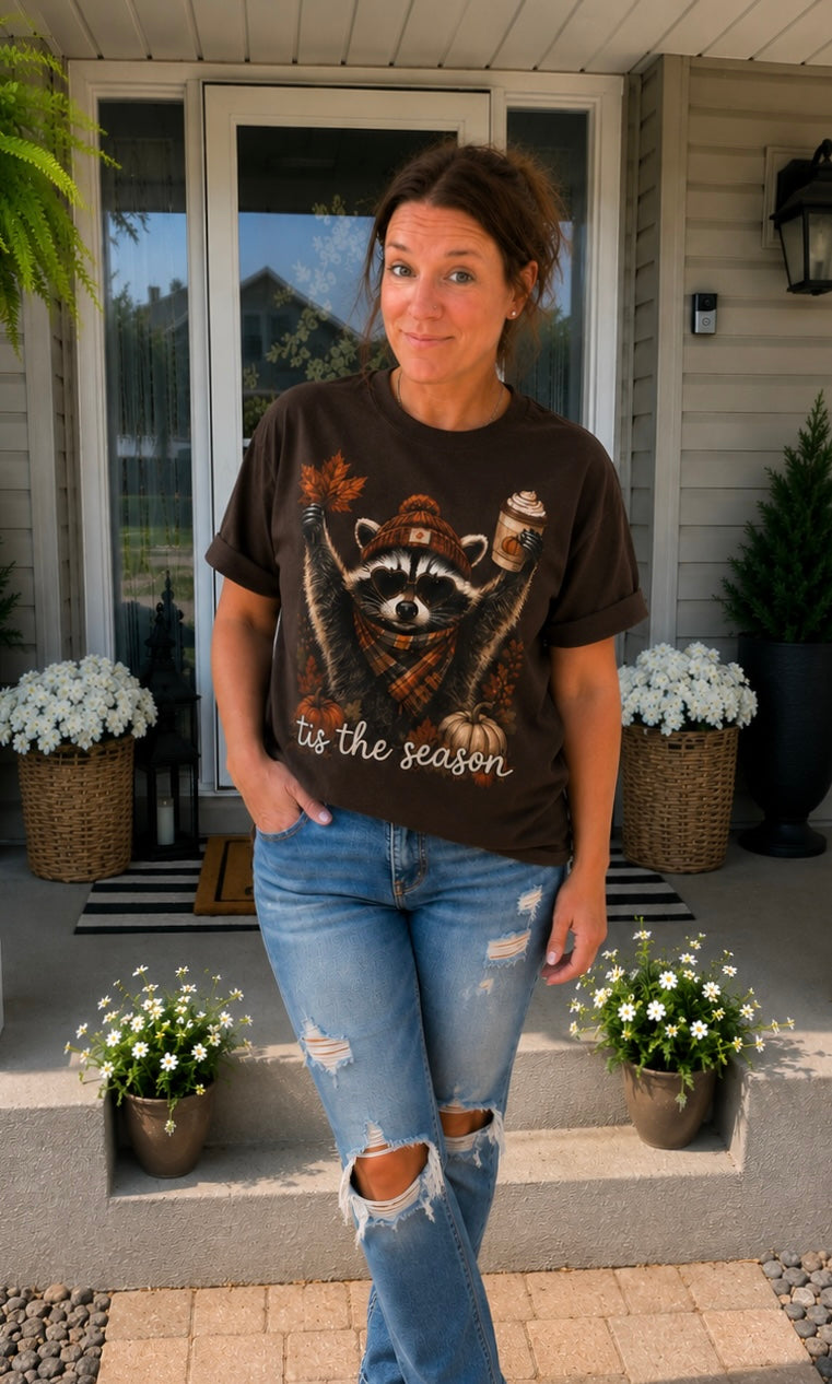 A woman stands on a front porch wearing a dark brown graphic t-shirt with a raccoon illustration and the text 'tis the season', paired with distressed blue jeans.