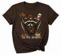 A dark brown short-sleeve t-shirt with a graphic of a raccoon wearing a knit beanie and scarf, holding a fall leaf and a coffee cup, with the text 'tis the season' and pumpkins below.
