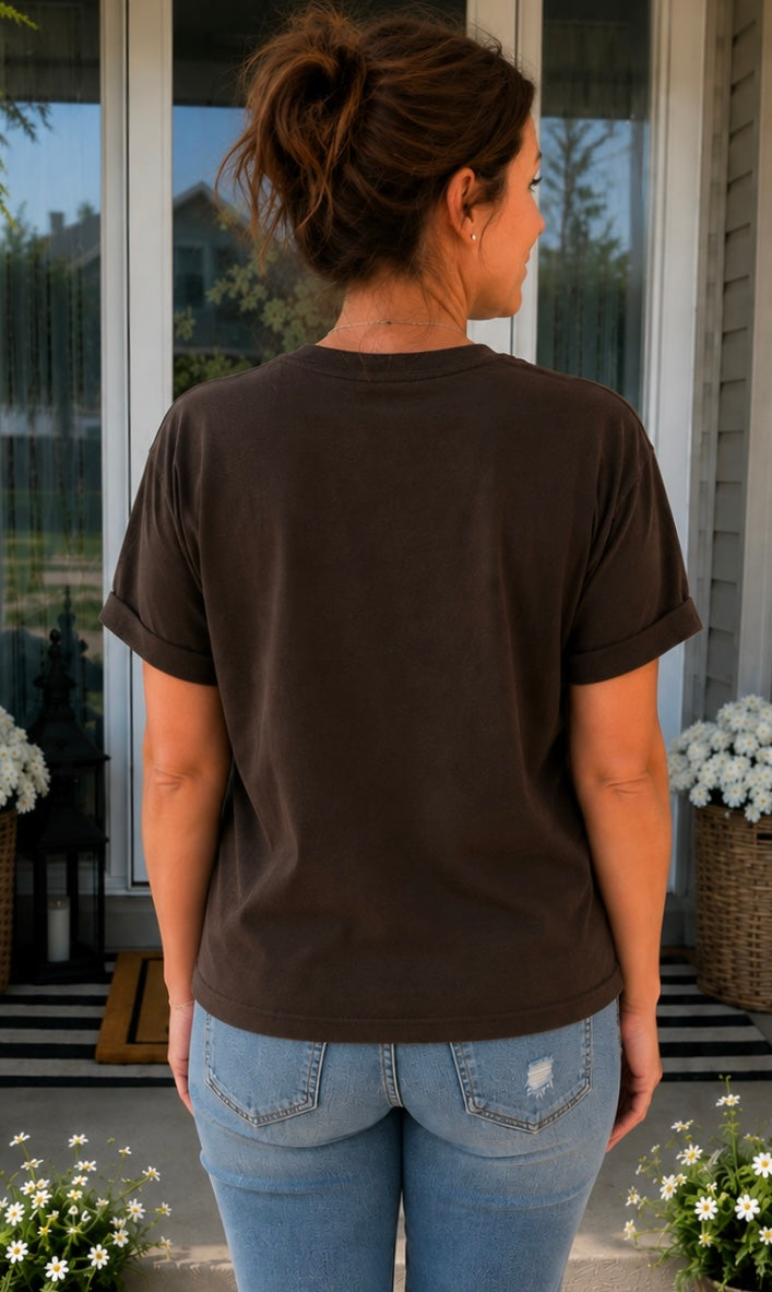 A woman viewed from behind wearing a dark brown short-sleeve t-shirt and light blue jeans, standing on a porch in front of a glass door.