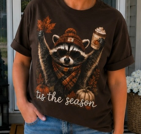 A person wearing a dark brown graphic t-shirt featuring a raccoon wearing a knit beanie and scarf, holding a coffee cup and autumn leaves, with the text 'tis the season' printed below.