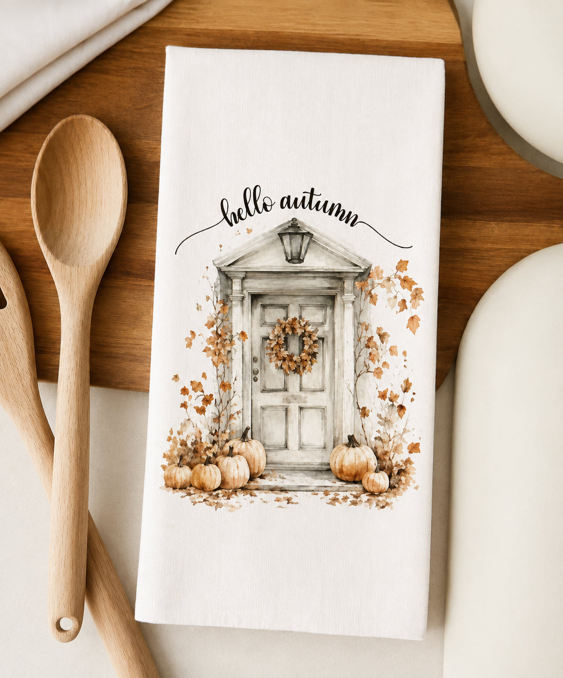 A white kitchen towel with a 'hello autumn' fall-themed illustration of a front door, pumpkins, and leaves, styled on a wooden board with wooden spoons.