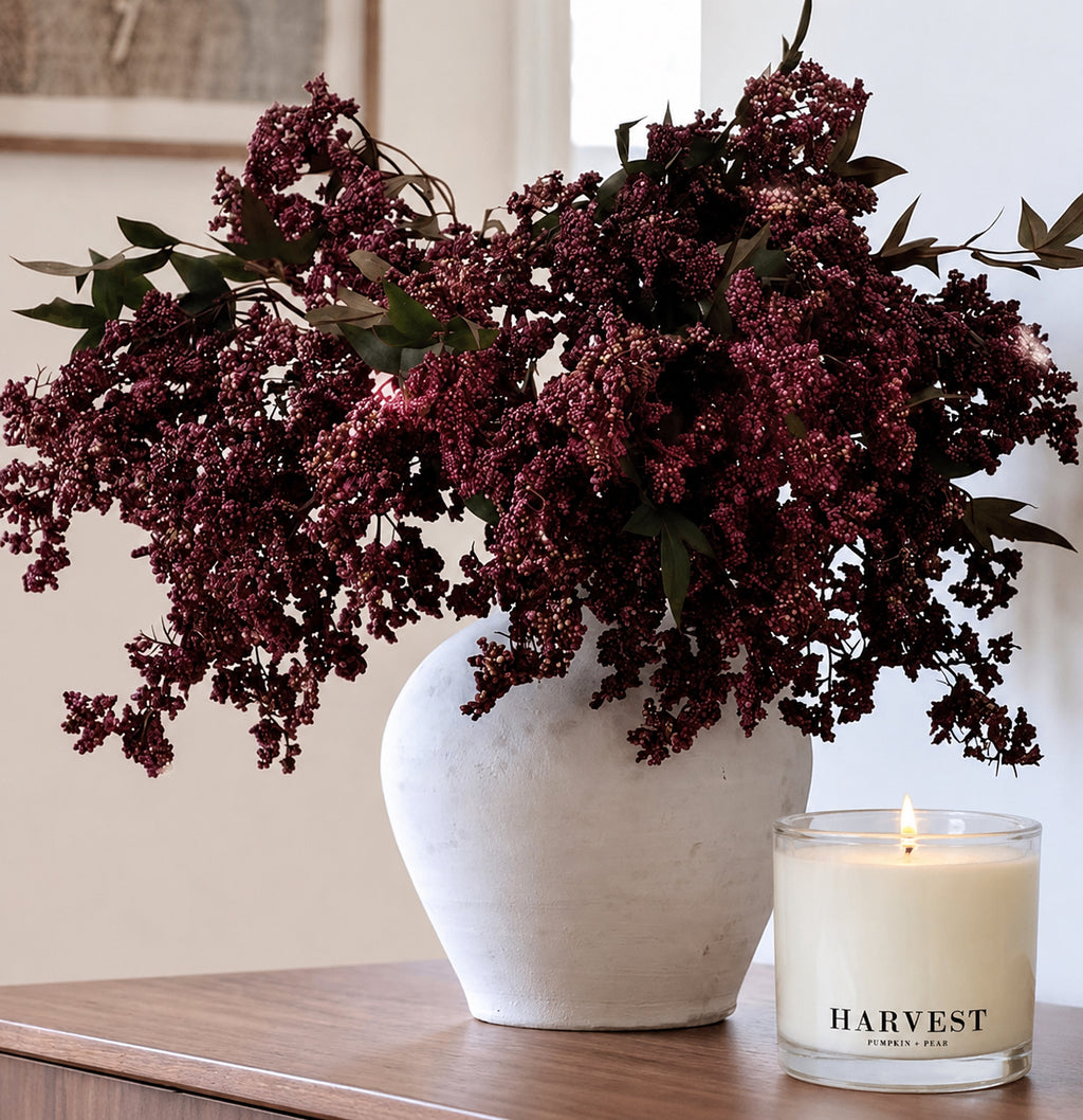A large white ceramic vase filled with dark burgundy dried flowers sits on a wooden table next to a lit candle in a glass jar labeled HARVEST.