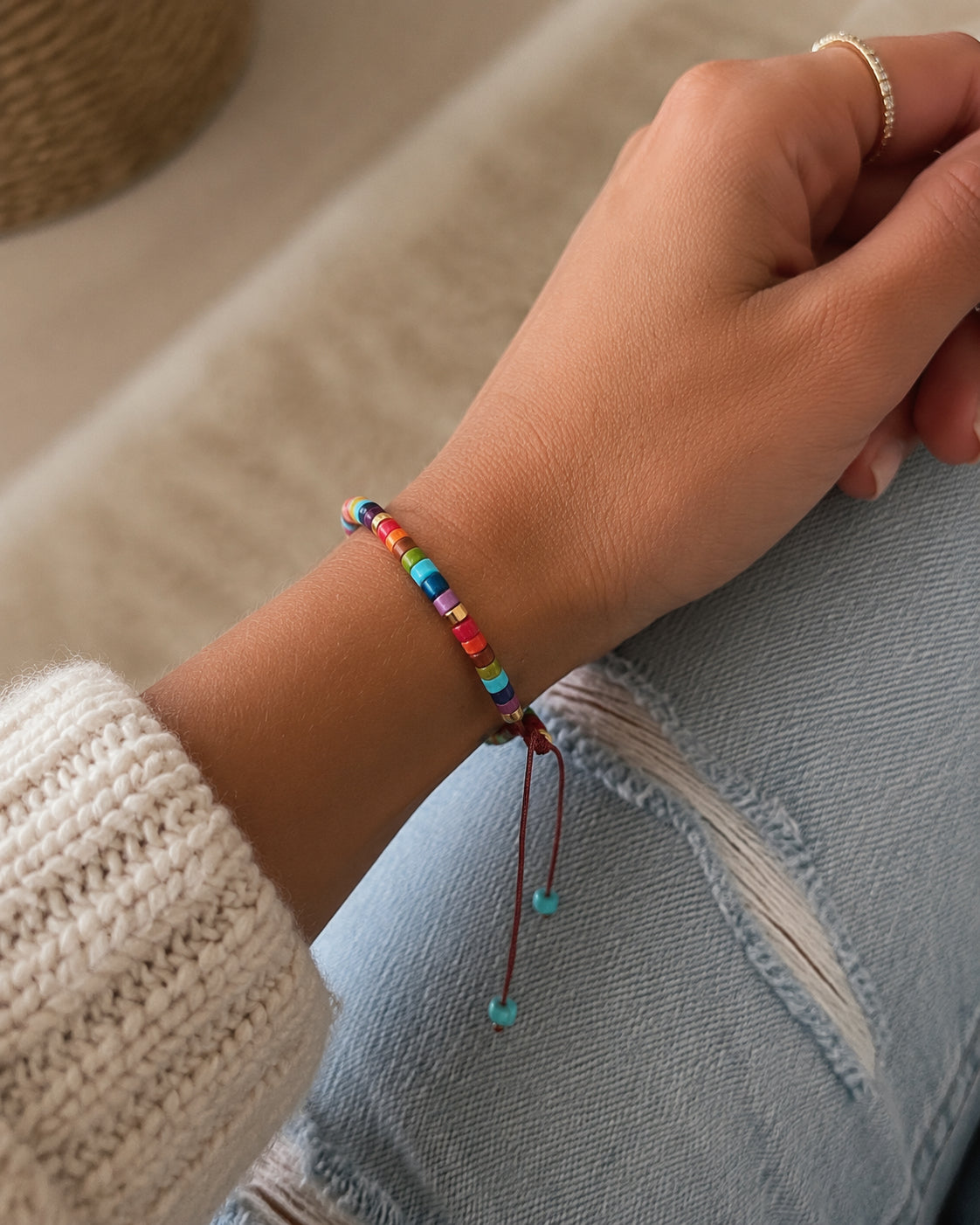 A close-up of a person's wrist wearing a colorful beaded cord bracelet with small turquoise beads, paired with a cream knit sweater and light blue jeans.