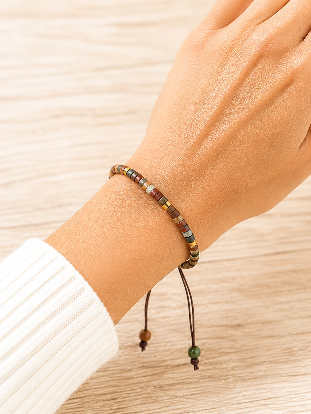A close-up of a person's wrist wearing a beaded adjustable cord bracelet, with a white ribbed sweater sleeve visible against a light wood background.